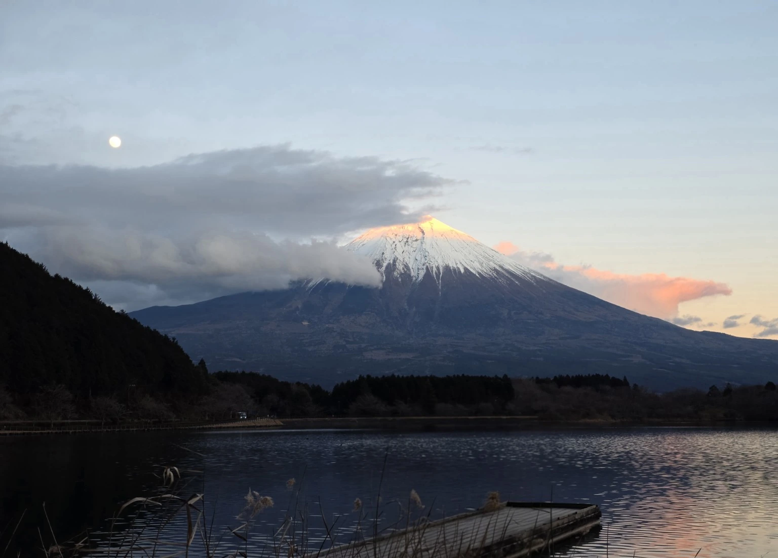 Mount Fuji above Lake Tanuki at sunset, its summit catching the last warm light as clouds wrap around the mountain and the moon hangs above