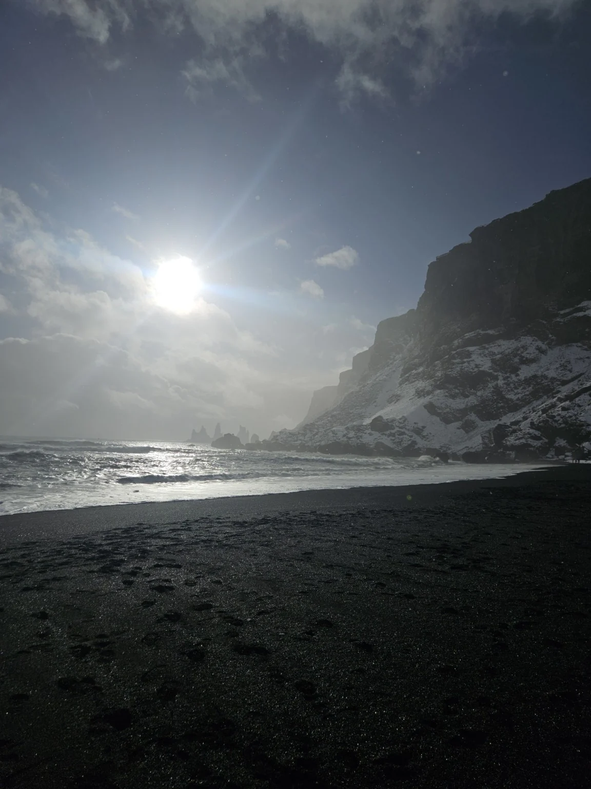 Sun breaking through winter cloud over Víkurfjara's dark sand, with Reynisdrangar sea stacks in the distance